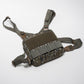 A tactical shoulder harness bag in a brown and green camouflage pattern, featuring a central hook-and-loop panel and adjustable straps, photographed on a light gray background.