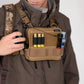 A close-up of a person wearing a brown jacket and camouflage cap, with a tan chest rig pouch holding shotgun shells and other gear against a plain light background.