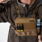 A close-up of a person wearing a brown tactical chest rig bag with magazine loops, and shotgun shell pouches, while holding a flashlight and wearing a brown jacket.