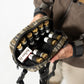 A person holds an open camouflage shotgun shell pouch containing multiple shotgun shells, with a shotgun magazine visible inside. The person is wearing a brown jacket and beige pants against a clean white background.