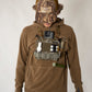 A man wearing a brown waffle-knit hoodie, khaki pants, and a brown camouflage cap stands against a plain light background. A camouflage chest rig loaded with cameras and accessories is worn over his hoodie.