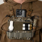 A close-up of a person wearing a brown hooded jacket and tactical chest rig with a camouflage utility pouch holding a smartphone, shotgun shell casings, a white bottle, and other gear.