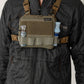 A man wearing a black puffer jacket and khaki pants models a tan chest rig bag with side bottle holders, shown from the neck down against a plain white studio background.