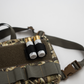 A close-up of a camouflage tactical pouch with olive green webbing and a brown plastic buckle, holding two shotgun shells in black loops.